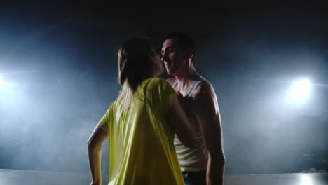 La-Cámara-Hace-Zoom-Y-Dispara-Desde-El-Plano-Completo-Al-Plano-Medio-En-Cámara-Lenta,-Una-Joven-Con-Un-Vestido-Amarillo-Y-Un-Hombre-Con-Una-Camisa-Blanca-Bailando-En-El-Escenario-Con-Focos-Y-Humo